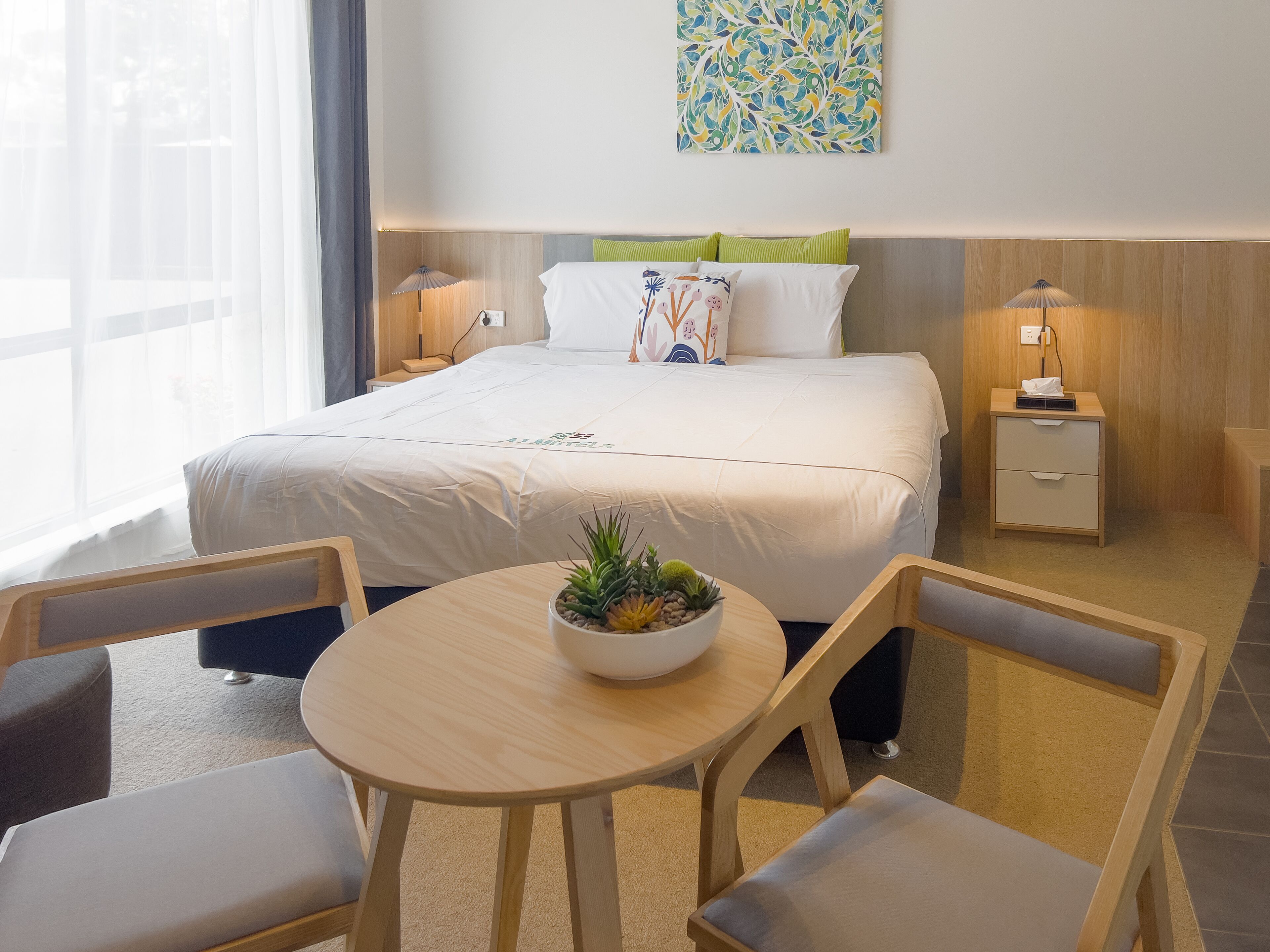 A hotel room with a bed, bedside tables, a small table with a plant, and two chairs.