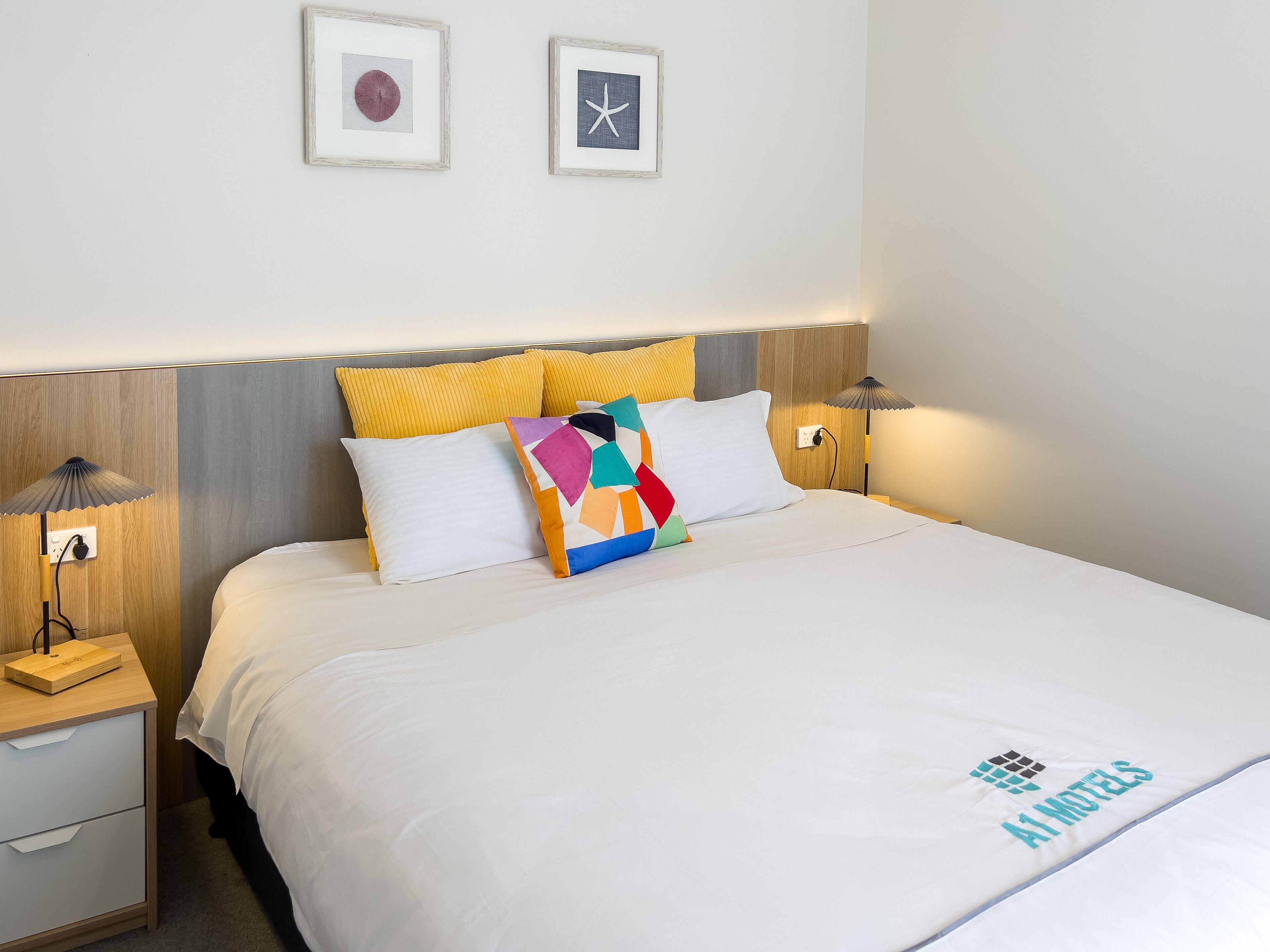 A neatly made bed with a white comforter and a colorful geometric pillow. There are two bedside lamps, a wooden nightstand, and a wall-mounted lamp. Two framed pictures are hung above the bed.