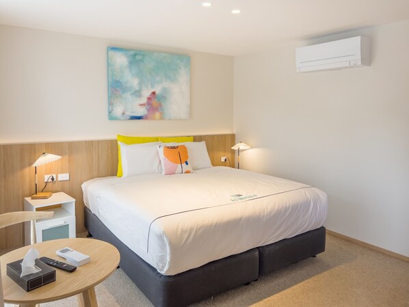 A hotel room with a bed, bedside lamps, a small table with a tissue box and remote, a painting on the wall, and an air conditioner.