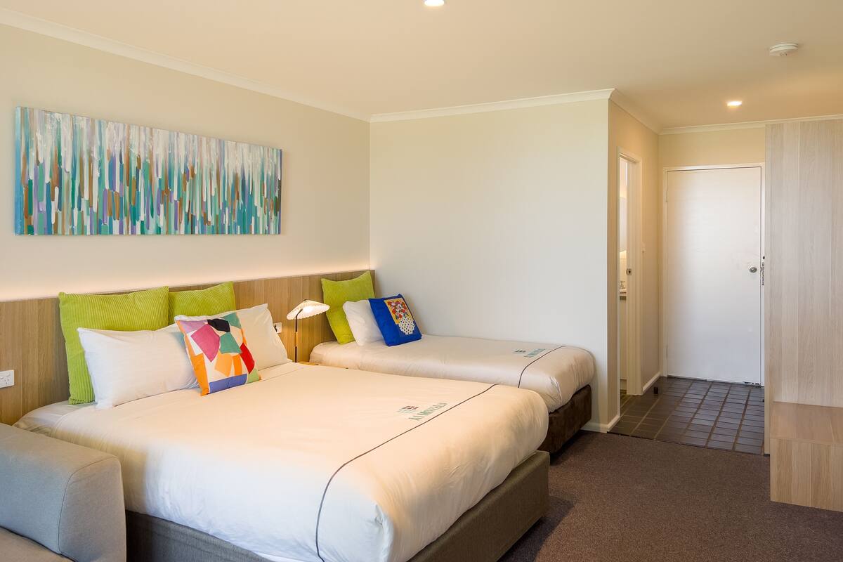 A hotel room with two beds, a large abstract painting, a wooden headboard, and a door leading to another room.