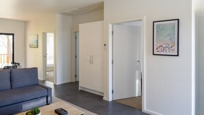 Una sala moderna con un sofá gris, una mesa de centro con una planta y un comedor con sillas. Se observa un armario blanco y una puerta que da a otra habitación.