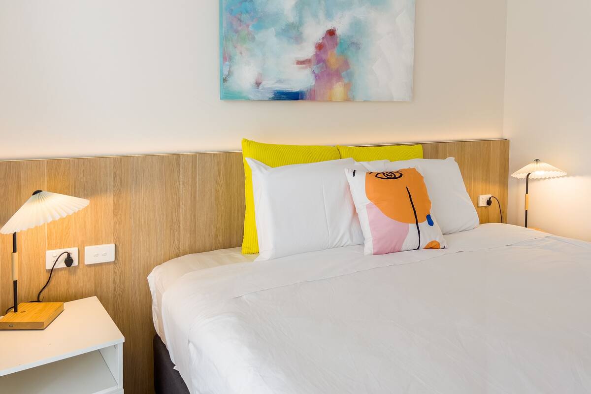A neatly made bed with white bedding, a wooden headboard, and a colorful abstract painting above it.