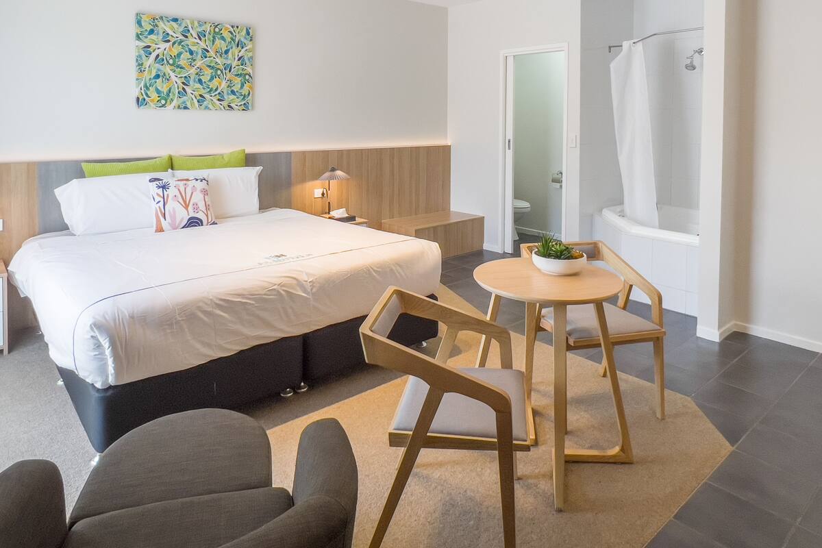 A modern hotel room with a large bed, a small table with chairs, a shower area, and a wall-mounted artwork.