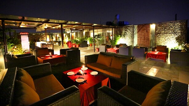 An outdoor dining area with wicker furniture and red tablecloths, featuring a Coca-Cola sign and potted plants. - Hotel Oyster (Chandigarh)