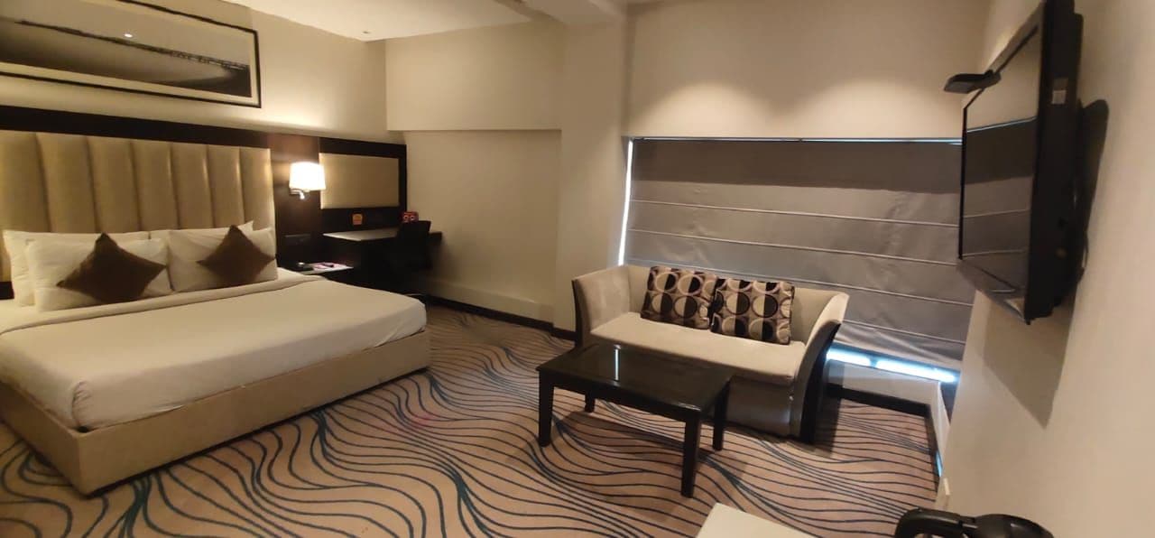 A hotel room with a bed, a sofa, a coffee table, a TV, and a patterned carpet.