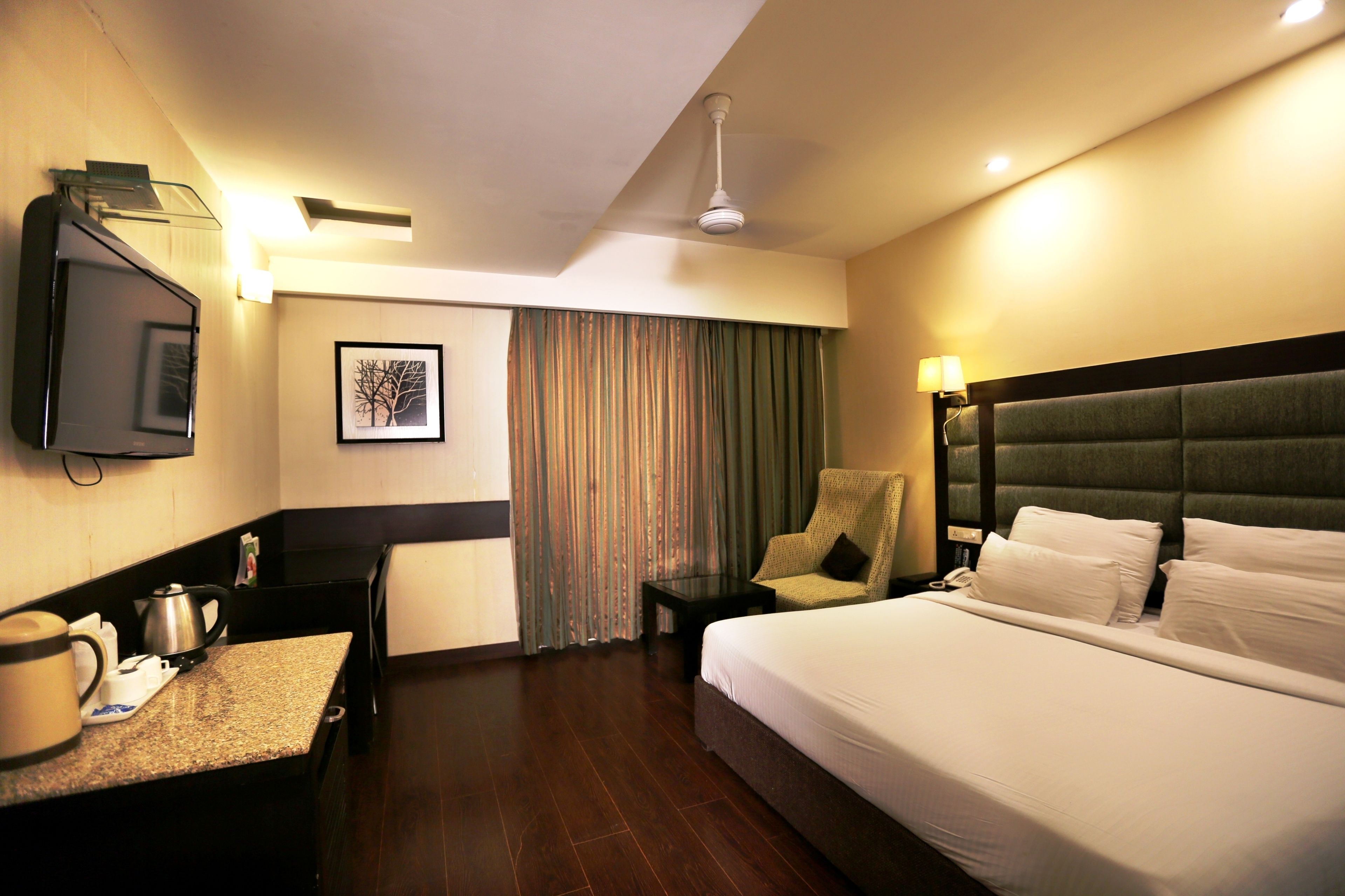 A hotel room with a bed, a desk with a kettle, a chair, a television, and a curtain.