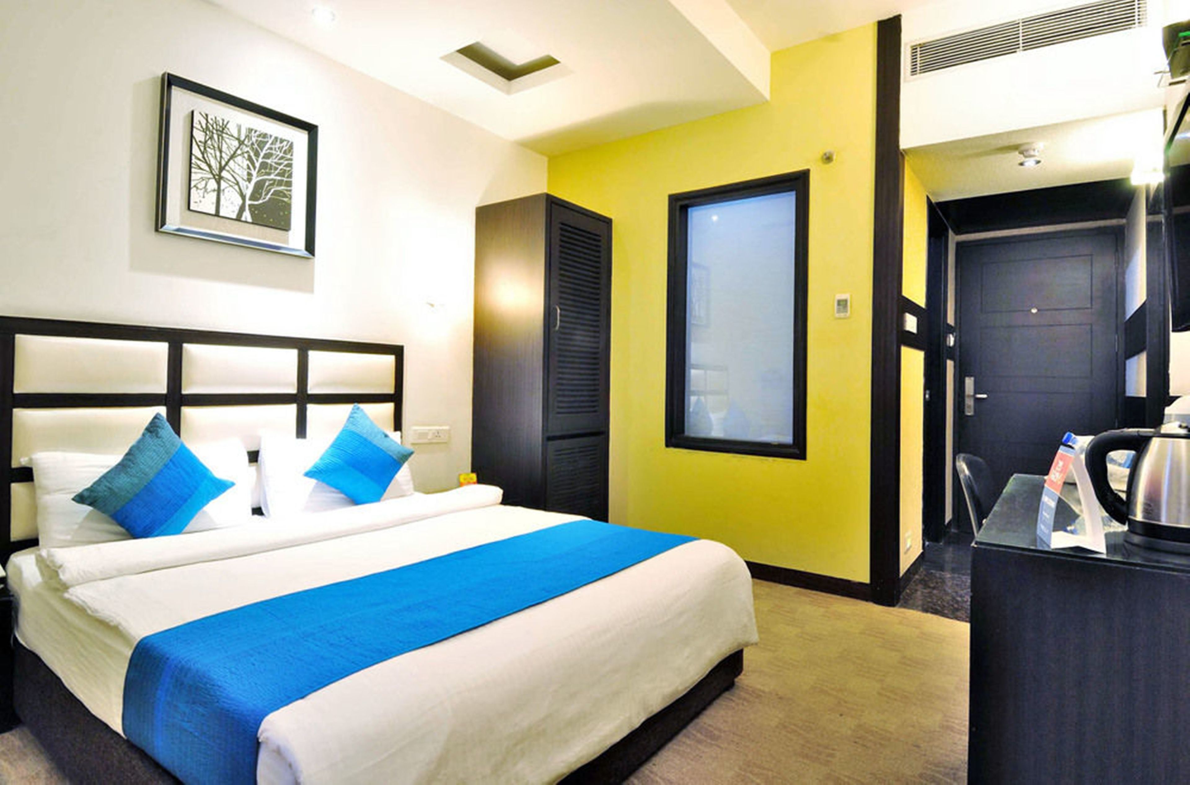 A hotel room with a bed, a desk with a kettle, and a door leading to a bathroom.