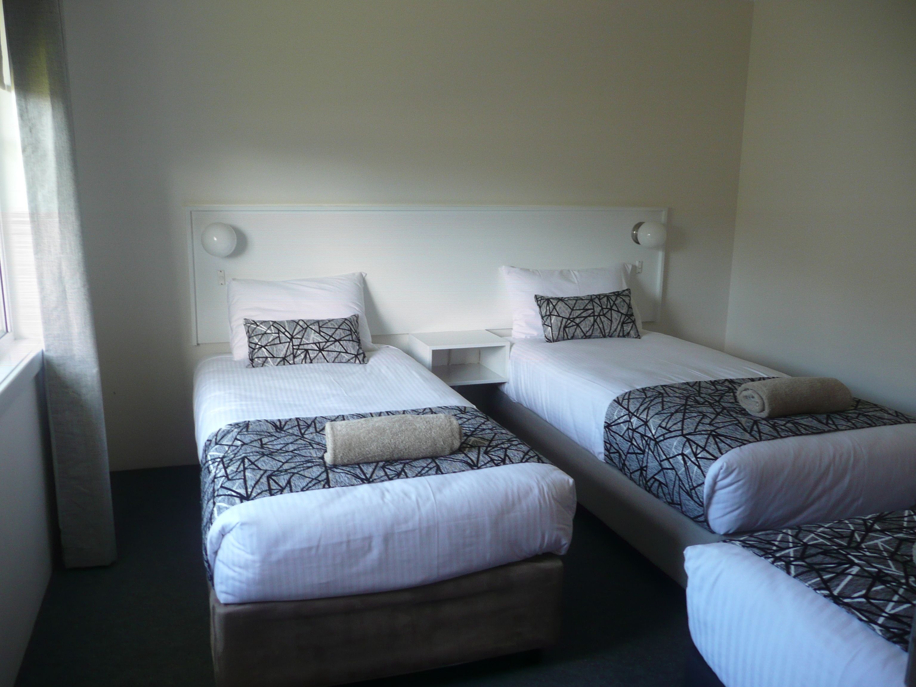 A hotel room with two single beds, each with a patterned bedspread and pillows, a bedside table, and a window with curtains.