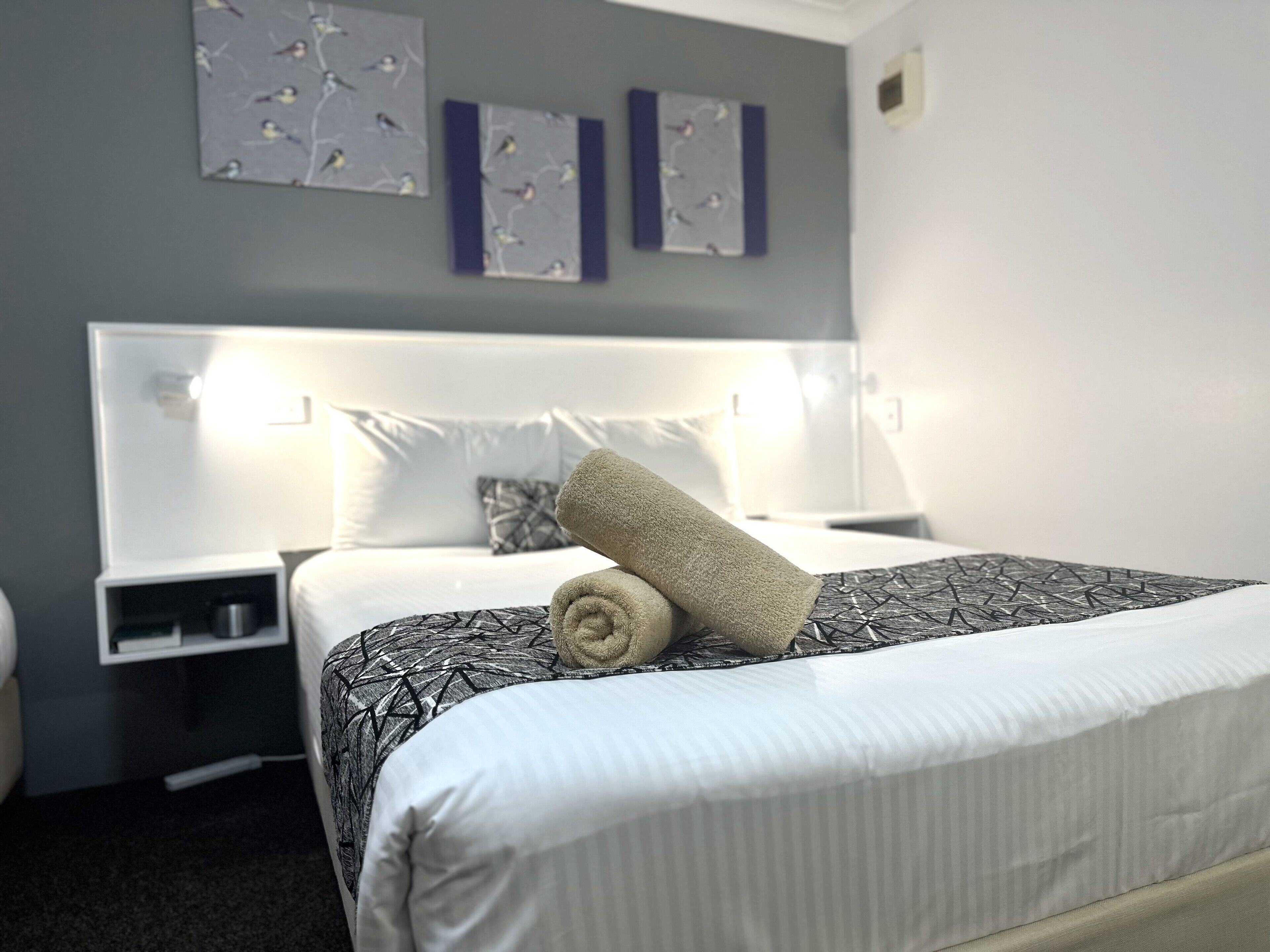 A neatly made bed with a rolled towel, a white headboard, and a bedside table with a lamp.