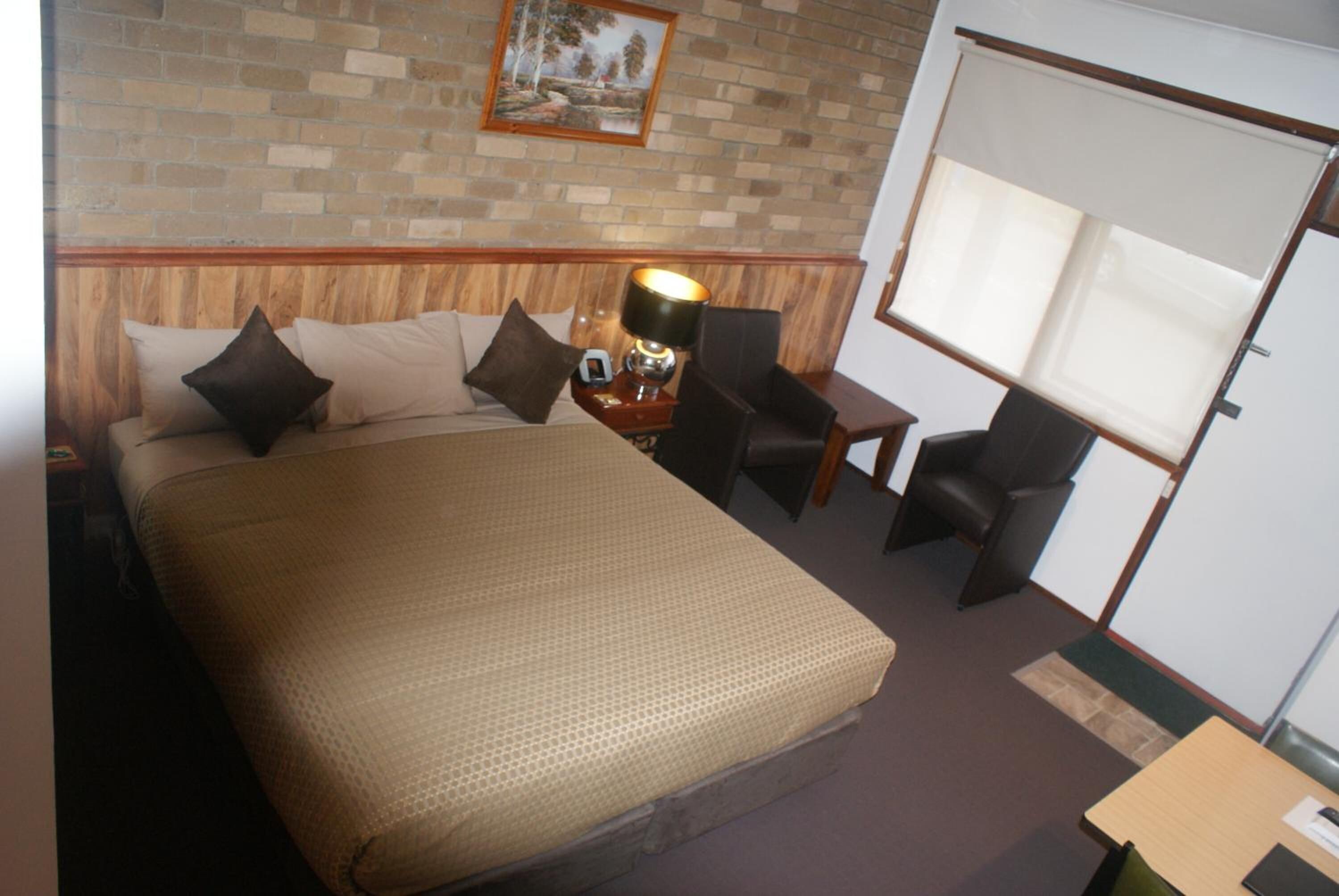 Een hotelkamer met een groot bed, twee stoelen, een tafeltje en een raam met jaloezieën.