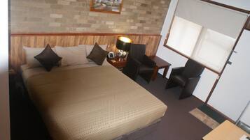 Een hotelkamer met een groot bed, twee stoelen, een tafeltje en een raam met jaloezieën.