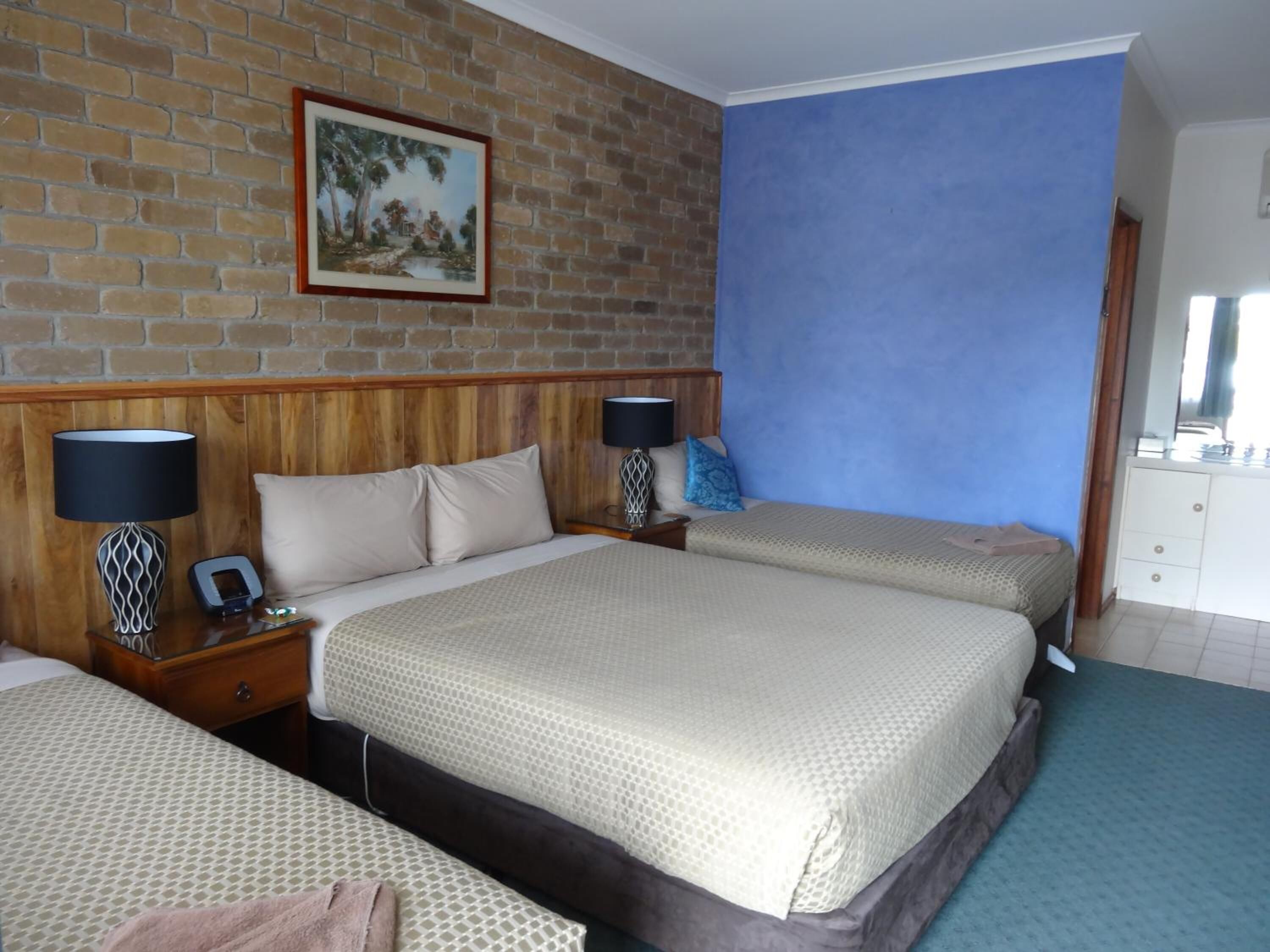 A hotel room with a brick wall, a blue accent wall, a wooden headboard, two bedside tables with lamps, a bed with a beige bedspread, and a small bedside table with a phone and a decorative item.