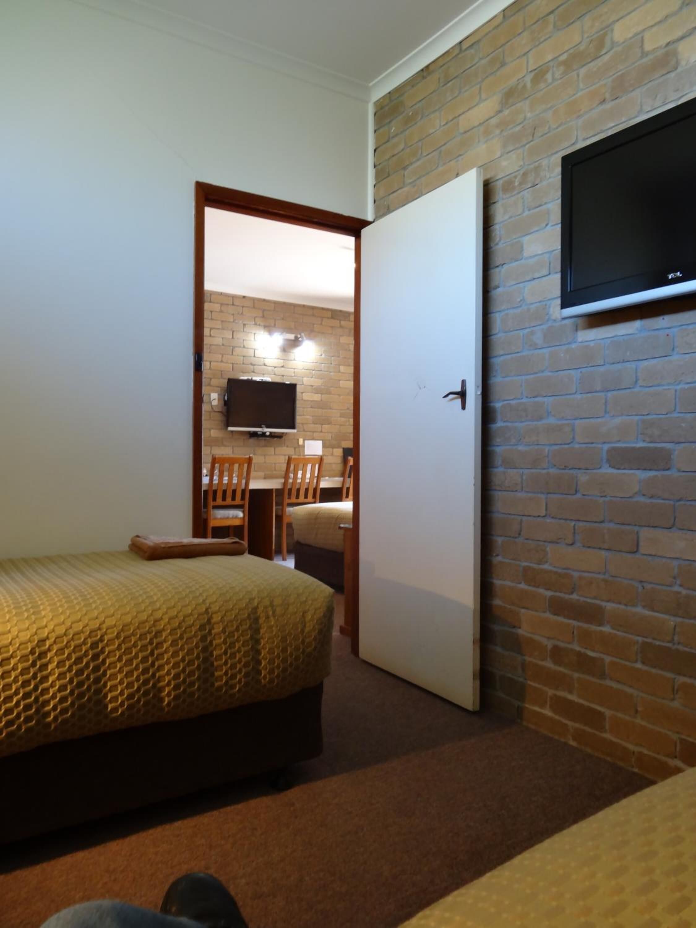 A hotel room with a bed, a TV mounted on a brick wall, a desk with a chair, and a door.
