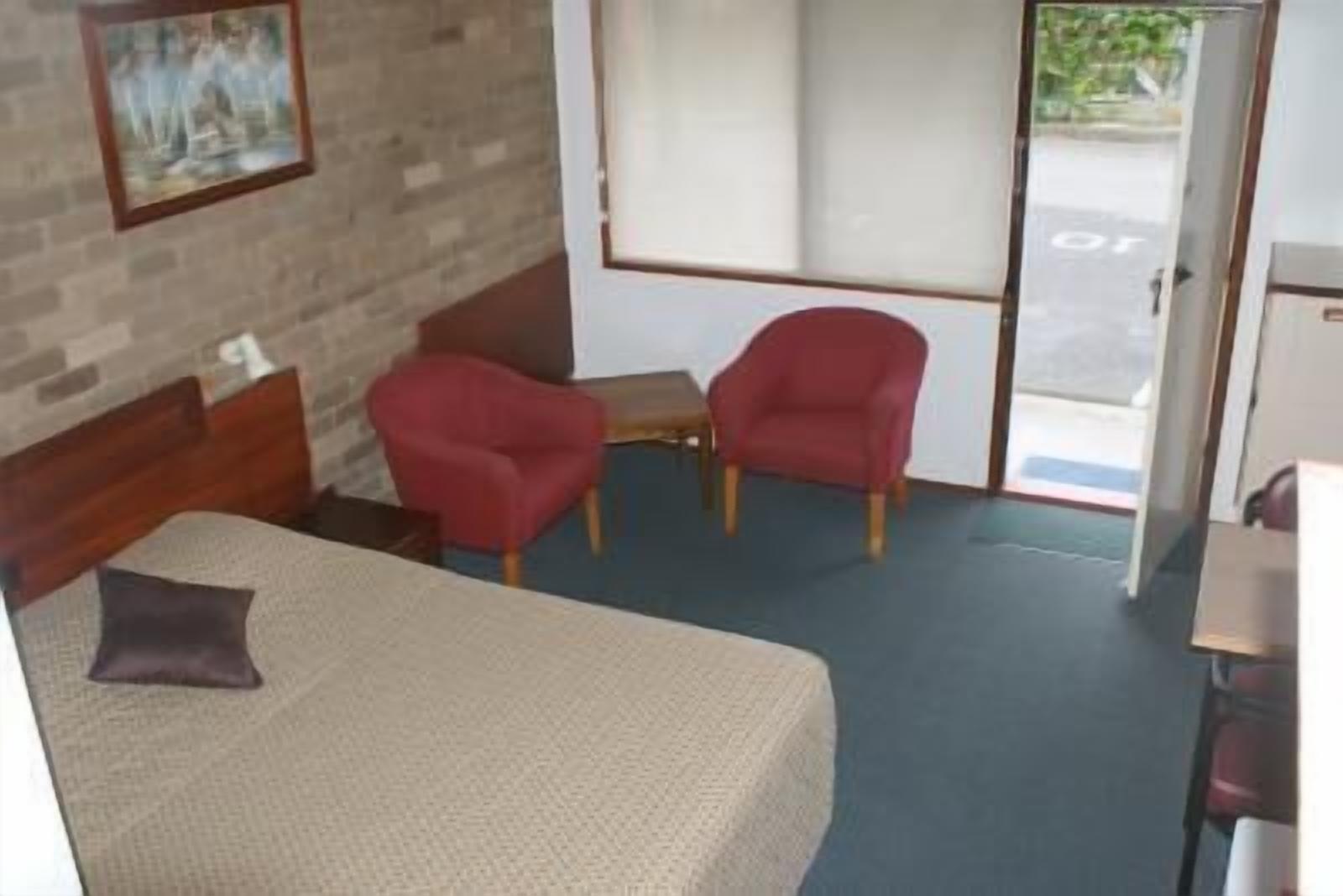 A hotel room with a bed, two red chairs, a small table, and a door leading outside.