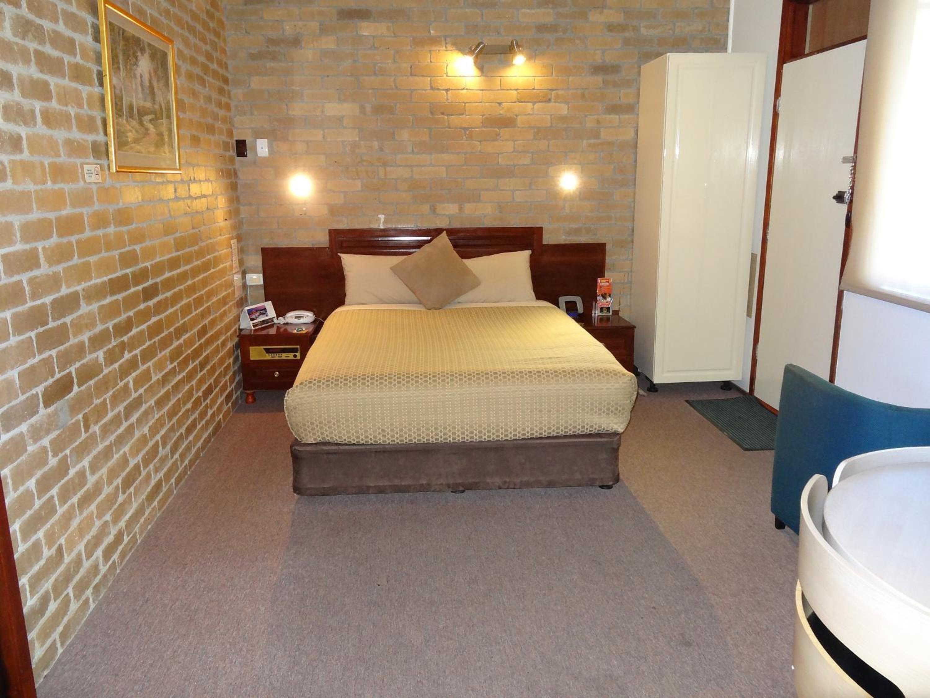 A hotel room with a brick wall, a wooden bed with a beige bedspread, a nightstand with a phone and a lamp, and a white wardrobe.