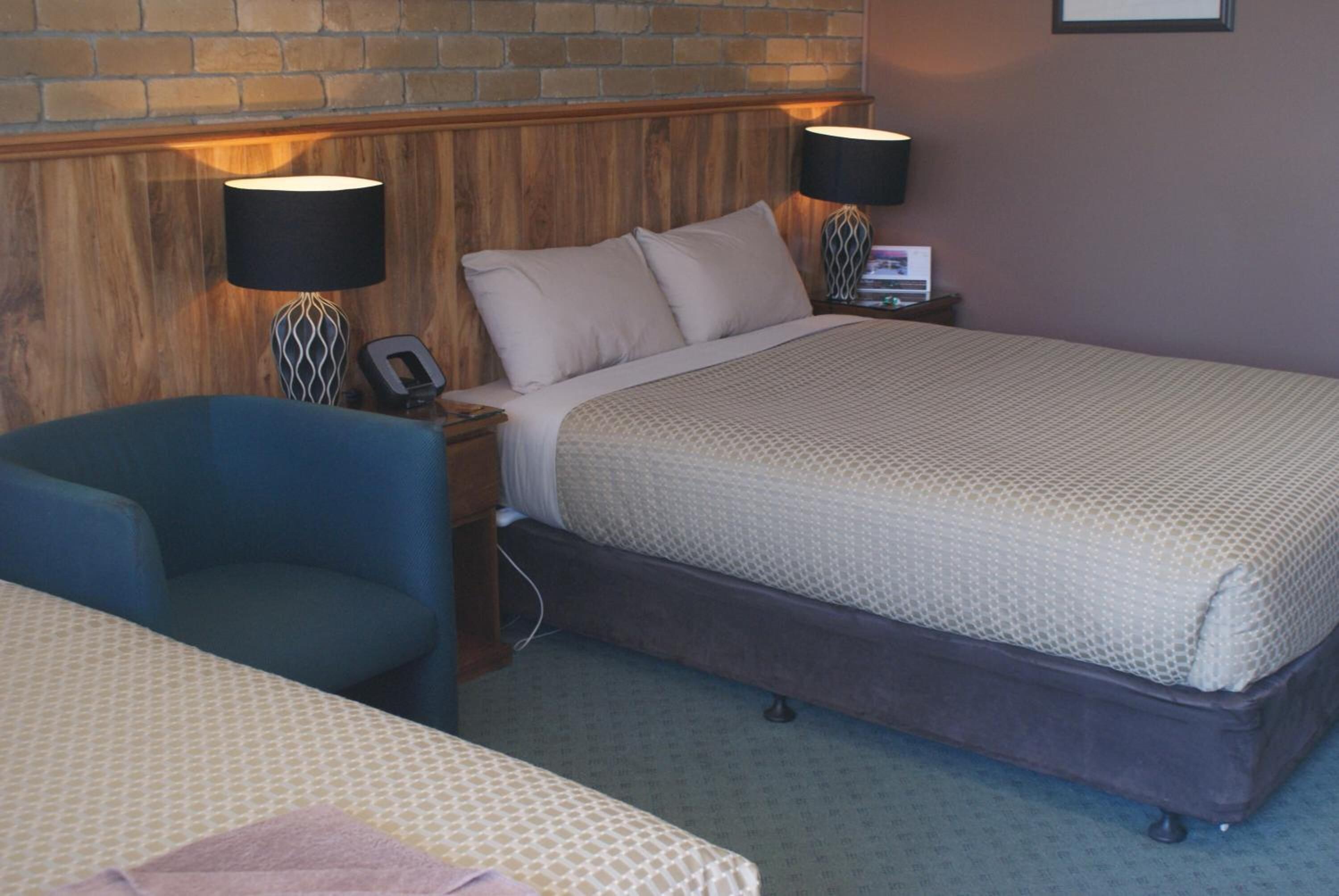 Une chambre d’hôtel avec un grand lit, deux lampes de chevet, une tête de lit en bois, un fauteuil bleu et une petite table sur laquelle se trouvent un téléphone et une lampe.