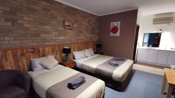 A hotel room with two beds, a wooden headboard, a brick wall, a painting, a mirror, and a kitchenette. - Peppinella Motel (Delacombe)