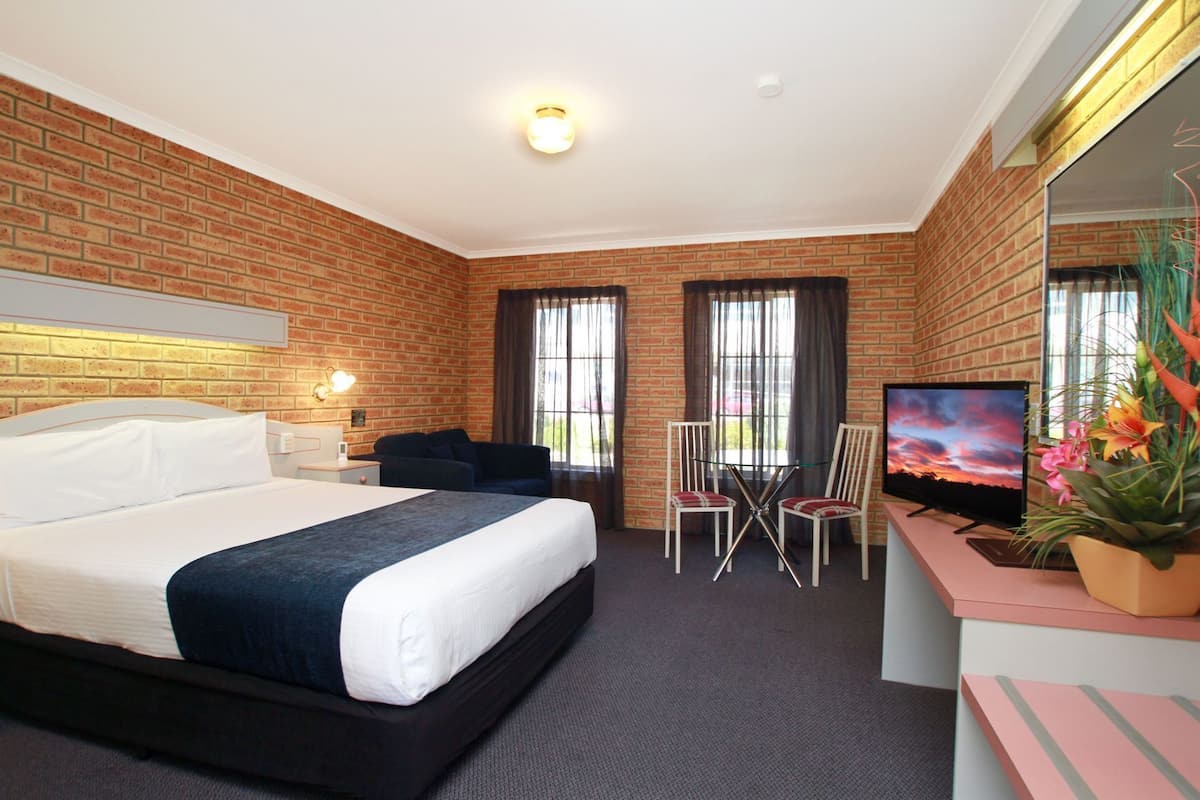 A hotel room with a brick wall, a bed, a desk with a TV, two chairs, and a large window.