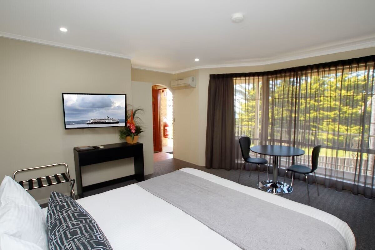 A hotel room with a bed, a TV mounted on the wall, a desk with a chair, a small table with chairs, and a large window with curtains.
