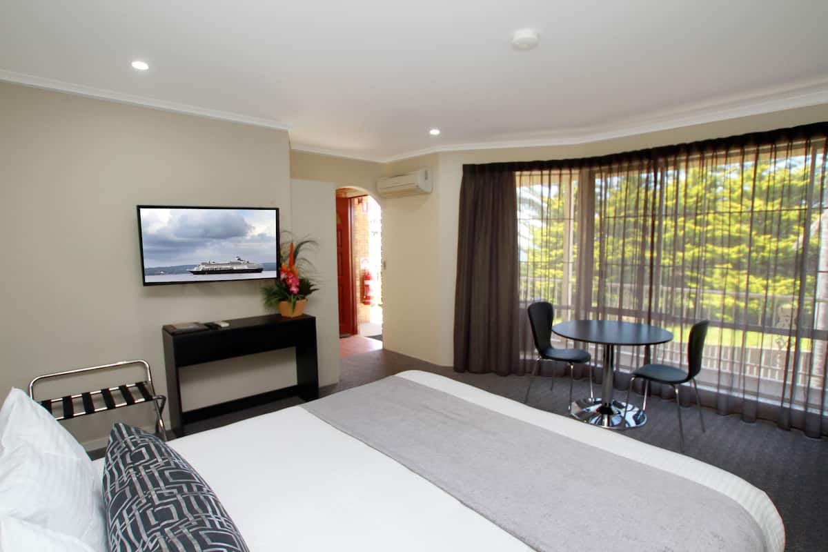 A hotel room with a bed, a TV mounted on the wall, a desk with a chair, a small table with chairs, and a large window with curtains.
