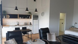 Una sala de estar moderna con sillas de cuero negro, una mesa de centro blanca y un sofá. Hay una zona de cocina con armarios blancos y un microondas.