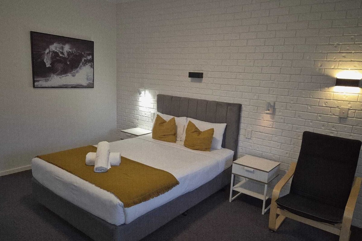 A hotel room with a bed, a chair, a nightstand, and a wall-mounted light.