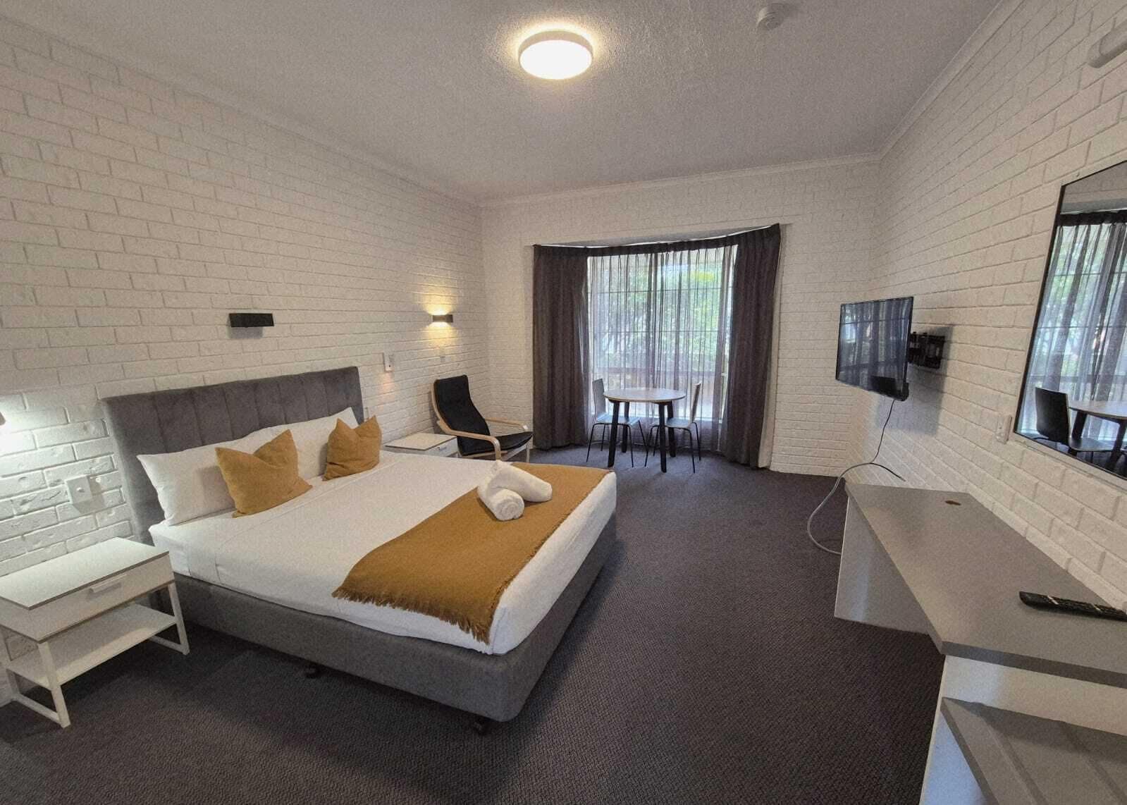 Ett hotellrum med en säng, ett skrivbord, en stol, en tv och ett fönster med gardiner.