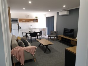 A modern living room with a sofa, coffee table, and TV. There is a dining area with a table and chairs, and a kitchen with a fridge and microwave.