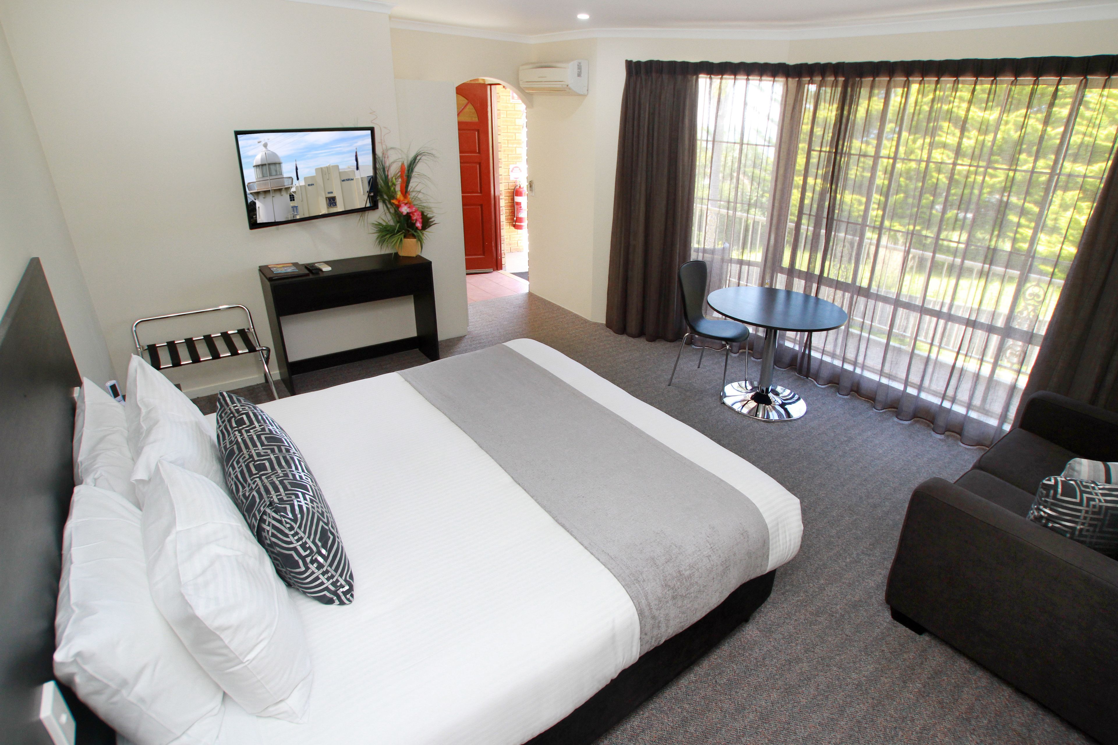 A hotel room with a large bed, a desk with a computer, a chair, a small table, and a balcony with a view of greenery.