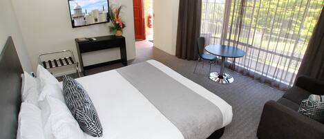 A hotel room with a large bed, a desk with a computer, a chair, a small table, and a balcony with a view of greenery.