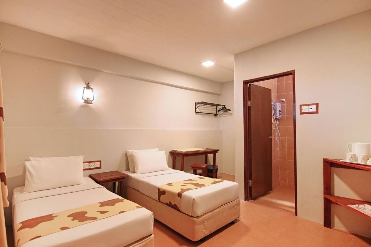 A hotel room with two beds, a small table, a chair, a desk, a shower area, and a towel rack.