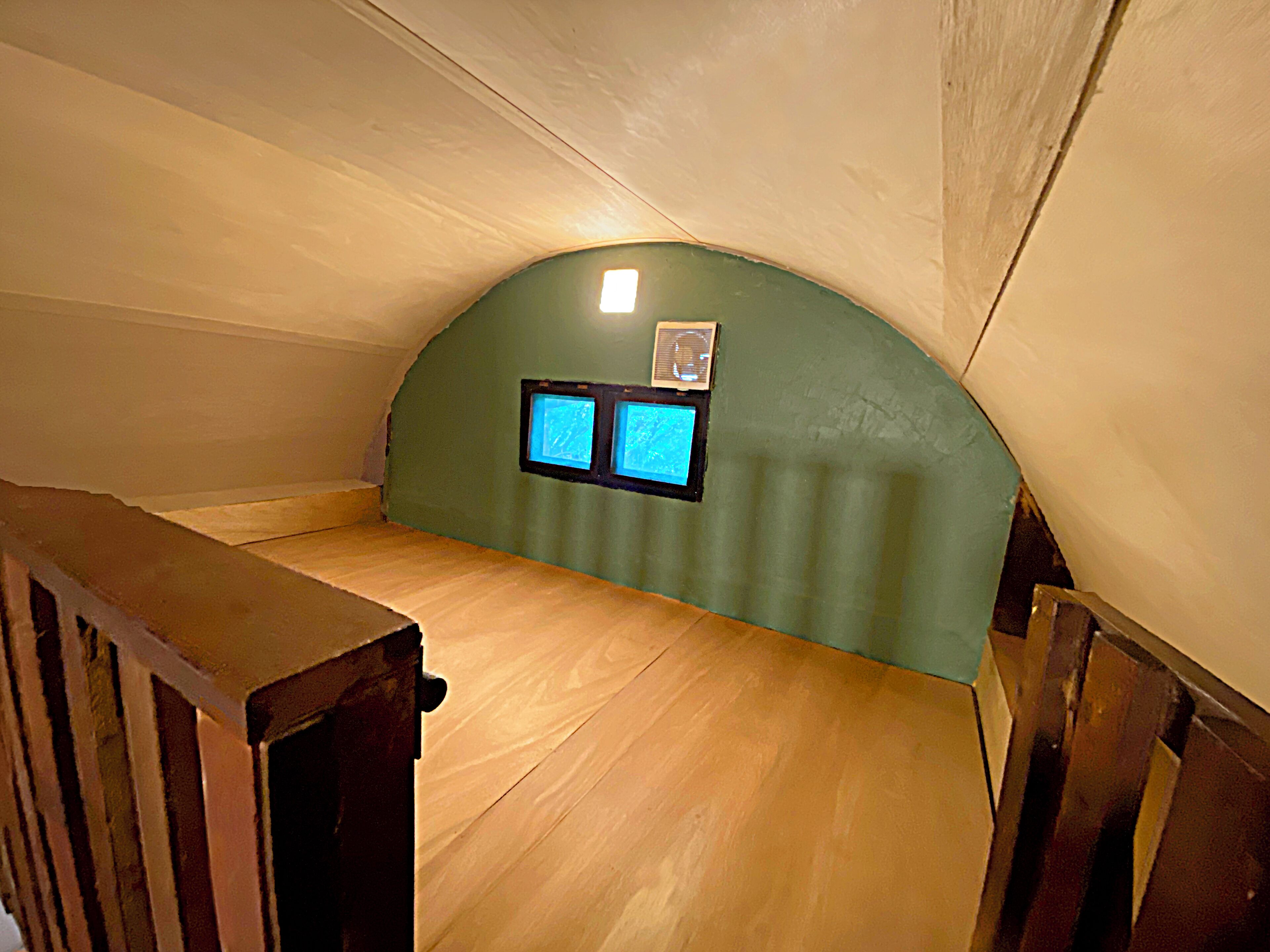 A small attic room with a sloped ceiling, a wooden bench, and two small windows with a view of the sky.
