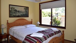 A neatly made bed with a striped blanket and a white pillow, a wooden headboard, a bedside table with a lamp, and a large window with a view of greenery and a brick wall. - Katoomba Townhouses (Katoomba)