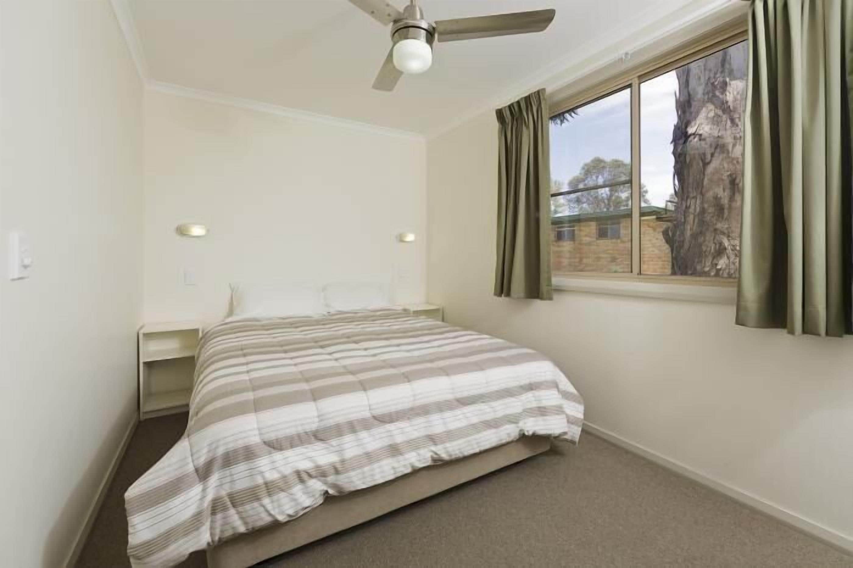 A bedroom with a bed, a ceiling fan, a window with curtains, and a view of trees and a building outside.