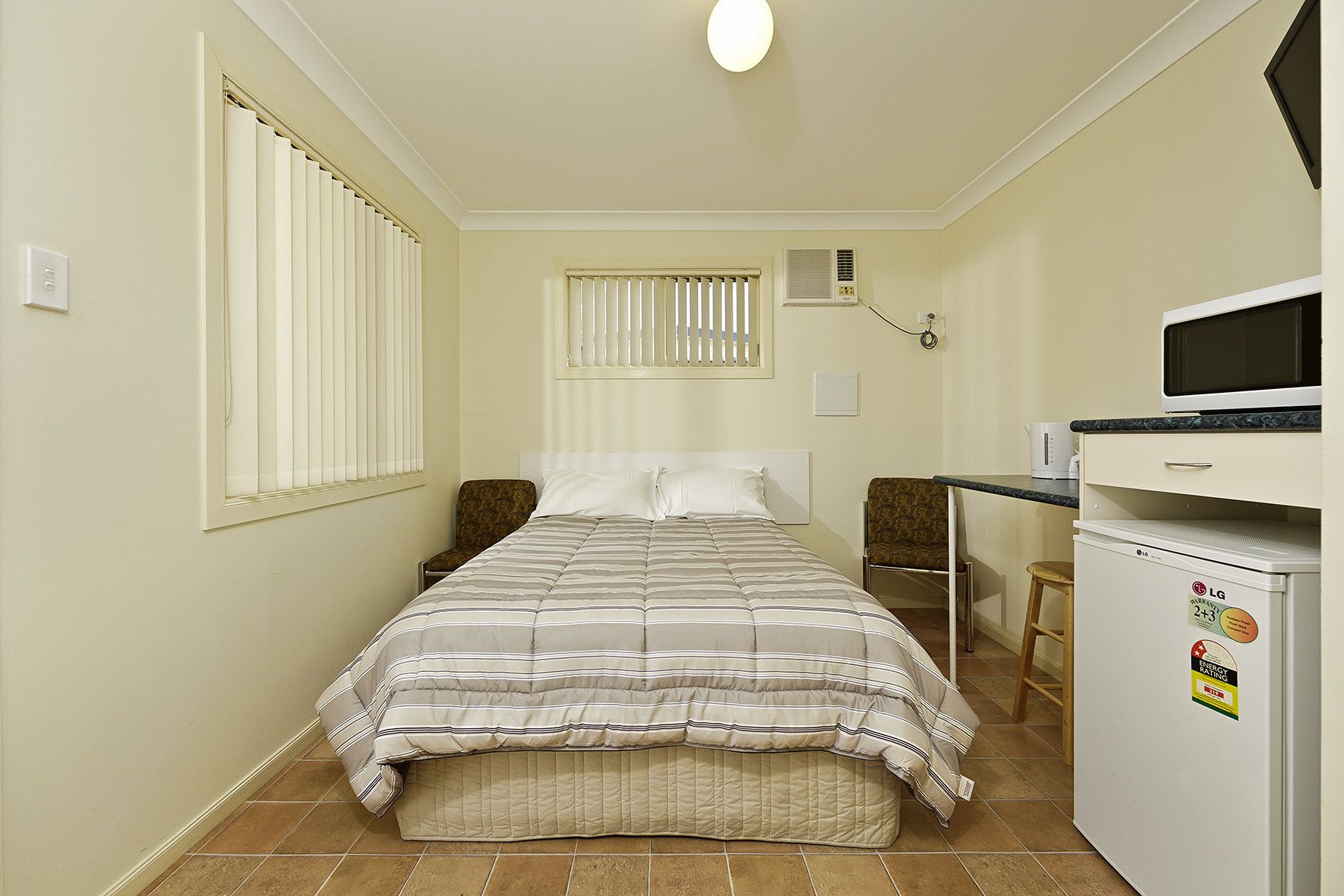 A compact hotel room with a bed, two chairs, a small kitchenette, and a window with blinds.