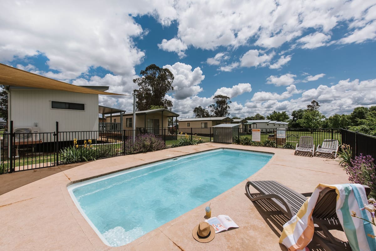 A swimming pool with lounge chairs and a covered area, surrounded by a fence and greenery.