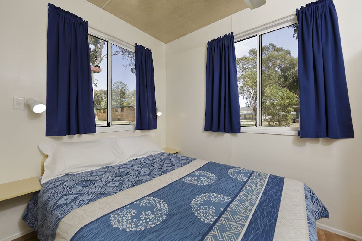 A bedroom with a bed, two windows with curtains, a bedside table, and a nightstand.