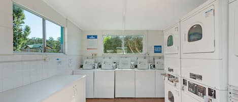 A laundry room with multiple washing machines and dryers, a window with a view of trees, and a tiled floor.