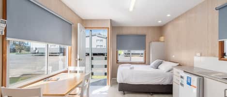 A compact hotel room with a bed, a small kitchenette, a dining area with a table and chairs, and a large window offering a view of the outside.