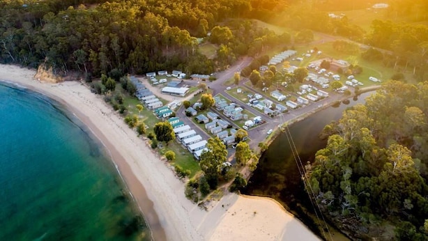 An aerial view of a coastal area with a beach, a row of caravans, and a forested hill in the background.
