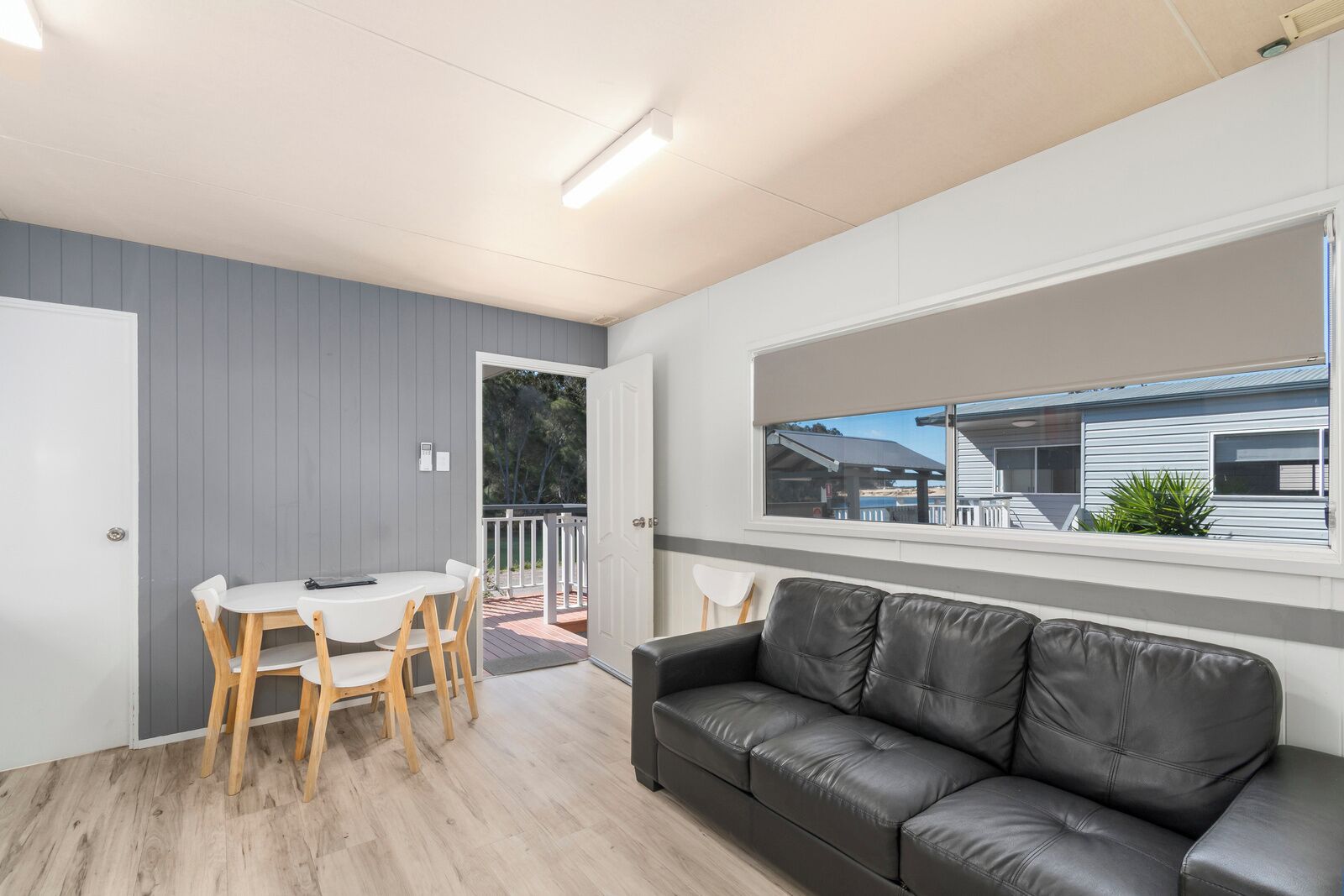 A modern living room with a black leather sofa, a white dining table with wooden chairs, and a large window offering a view of a neighboring house.