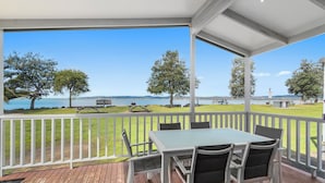 A covered outdoor area with a table and chairs, overlooking a grassy area with picnic tables and trees, and a view of the water.