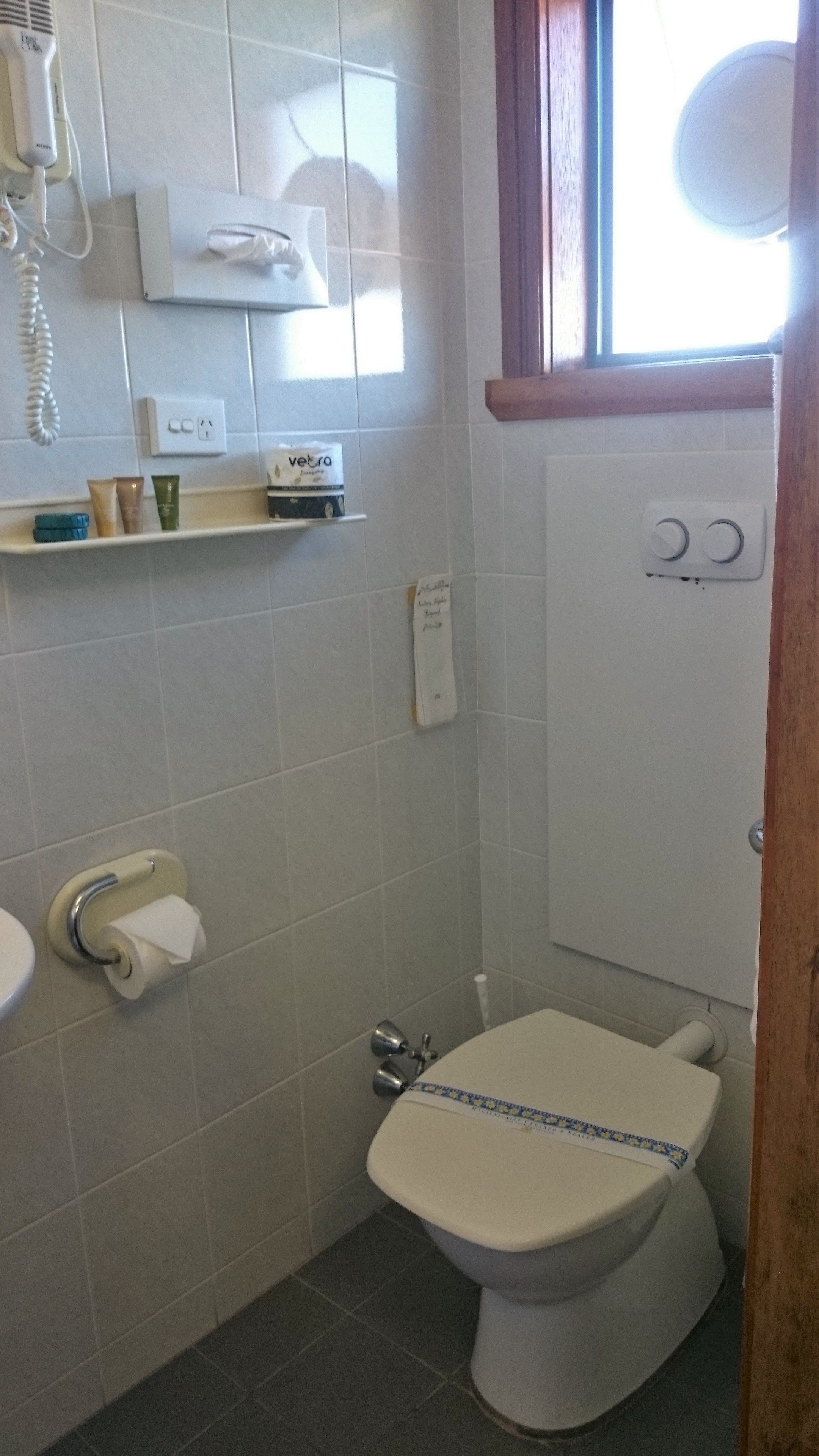 A small, compact bathroom with a toilet, a sink, and a shower area. There is a shelf with toiletries and a hairdryer on the wall.