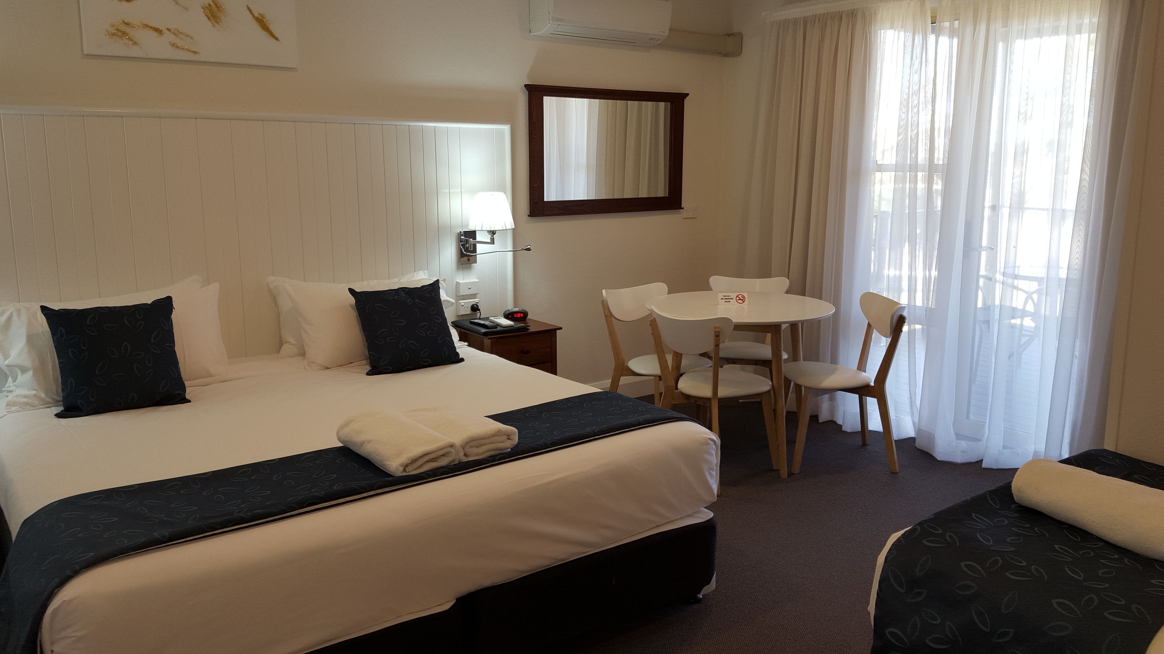 Quarto de hotel com cama, secretária, cadeira, mesinha e janela com cortinas.