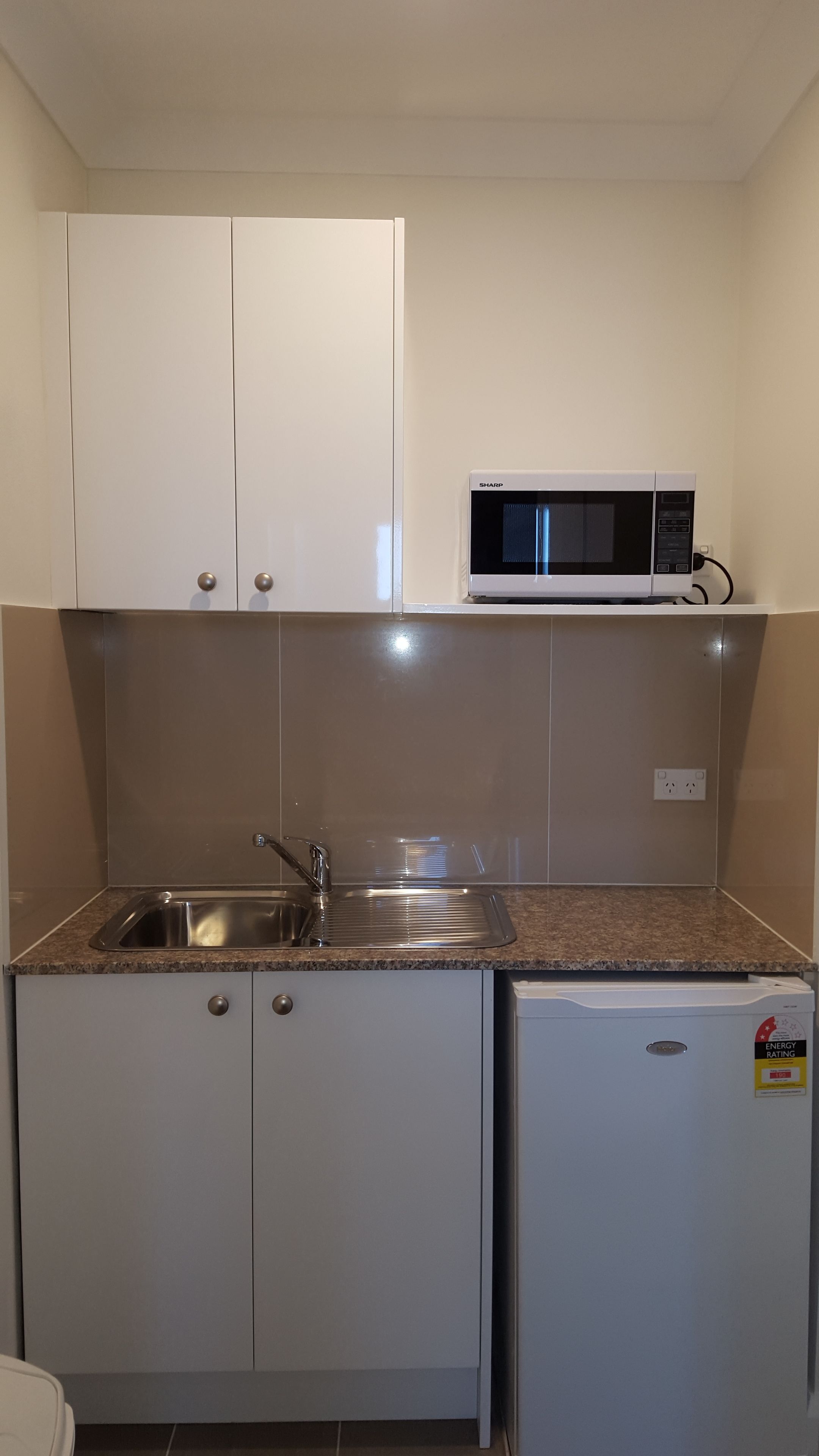 Een kleine keuken met witte kastjes, een roestvrijstalen spoelbak, een magnetron en een kleine koelkast.