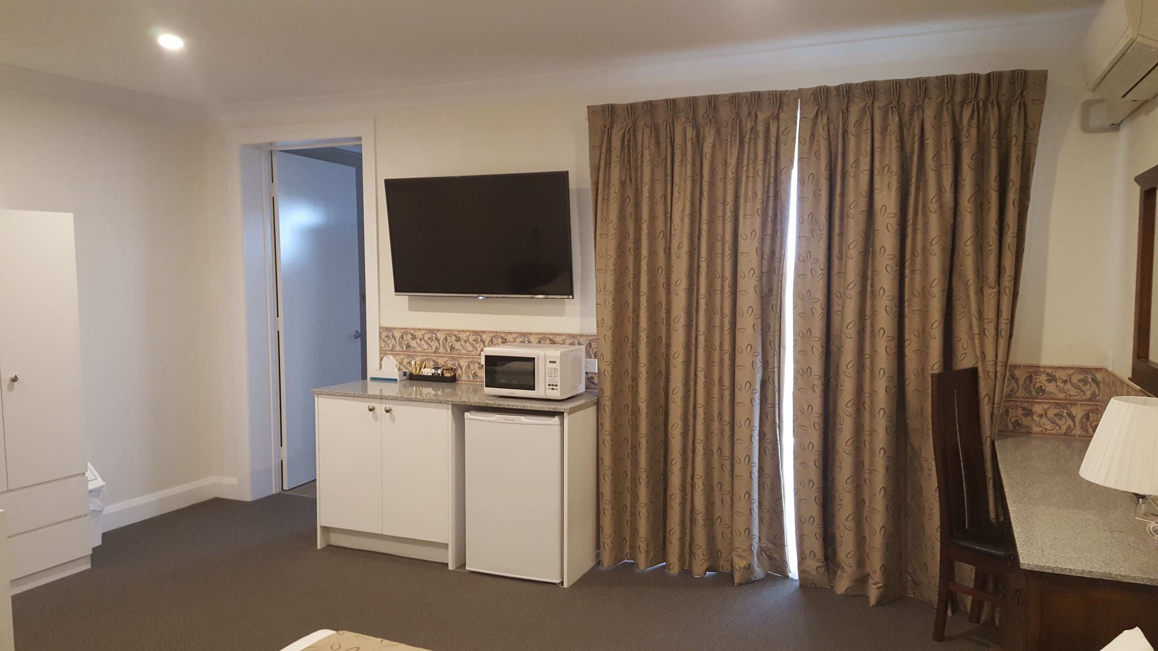 A hotel room with a flat-screen TV mounted above a kitchenette, a microwave, and a refrigerator. There is a door leading to another room, and a window with curtains.