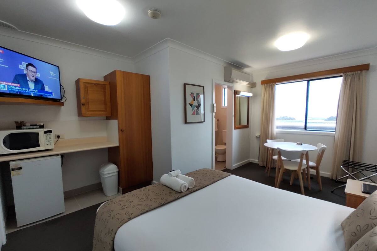 Habitación de hotel con cama, escritorio con televisor, refrigerador pequeño, microondas, baño y ventana con vistas.