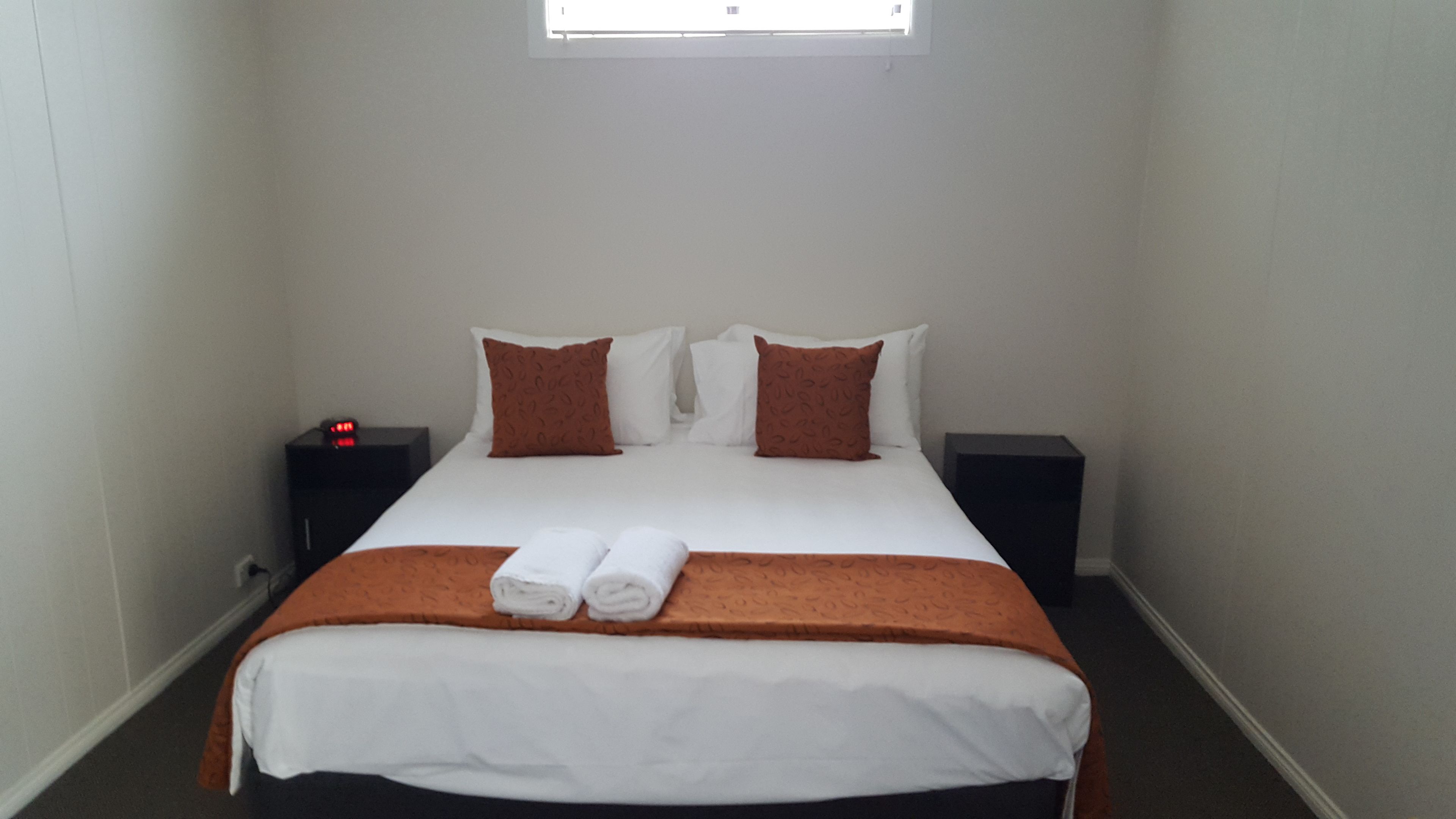 Een netjes opgemaakt bed met wit en bruin beddengoed, twee nachtkastjes en een klein raam boven het bed.