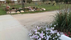 Ein Wohneingang mit einem Betonweg, der zu einem verschlossenen Eingang führt. Blumenbeete mit weißen und rosa Blüten, eine hölzerne Pergola und ein Spielplatz mit Spielgeräten im Hintergrund sind zu sehen.