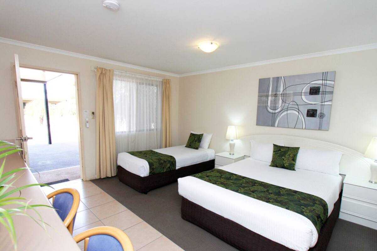 A hotel room with two beds, a desk, chairs, a window with curtains, a painting on the wall, and a door leading outside.
